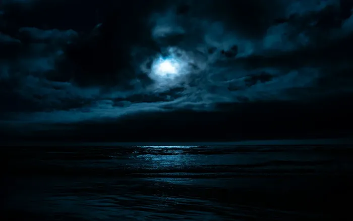 A photograph of a body of water illuminated by a full moon that's partially obscured behind some clouds