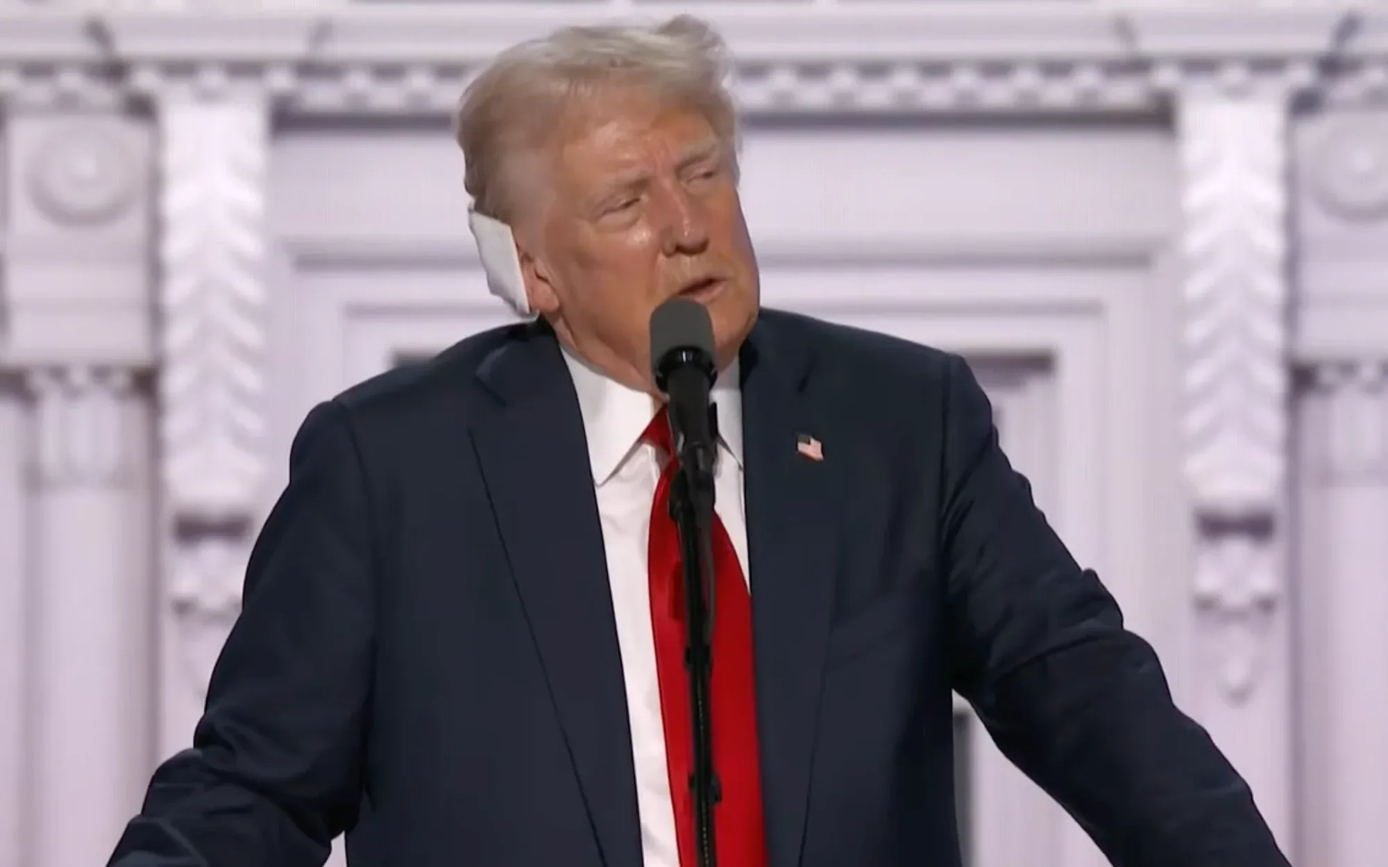 Donald Trump giving a speech in a blue suit and red tie, with the white bandage on his right ear