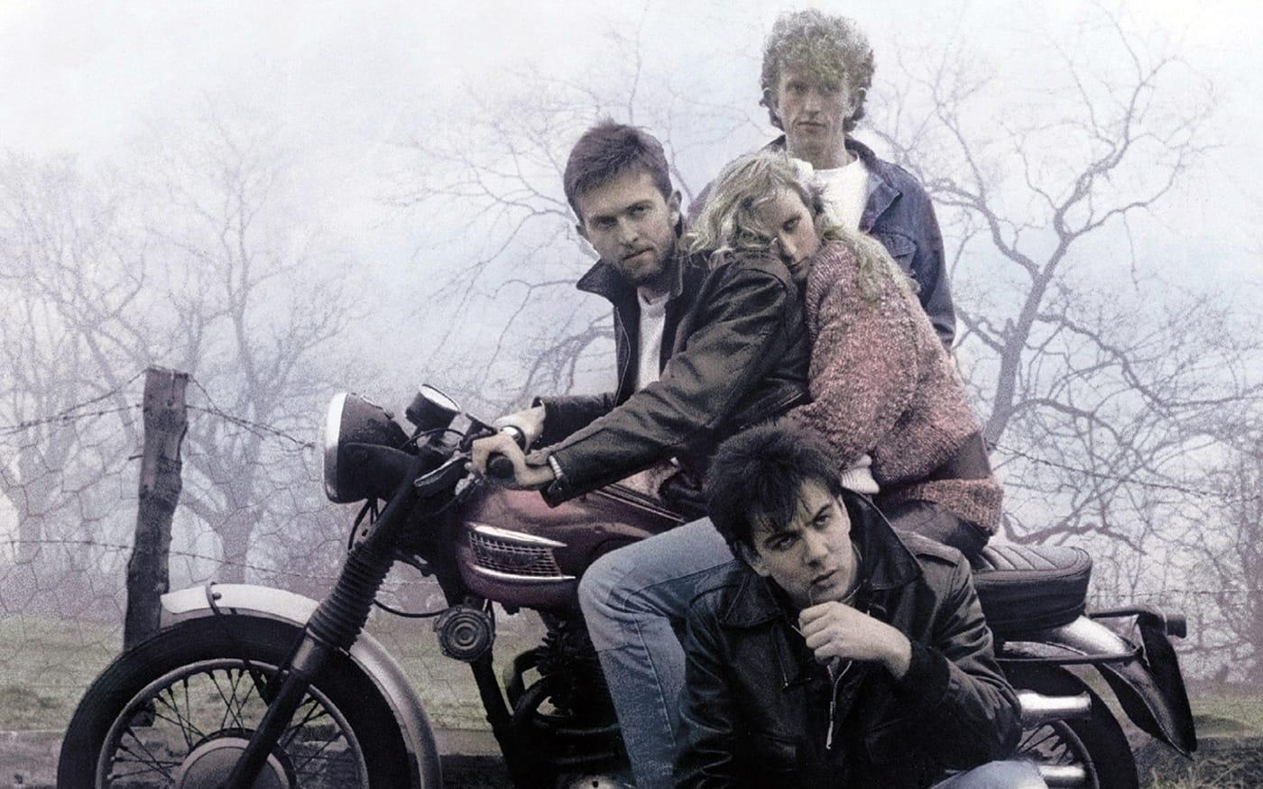 A photo of Prefab Sprout in a foggy countryside with two of the members sitting on a motorcycle