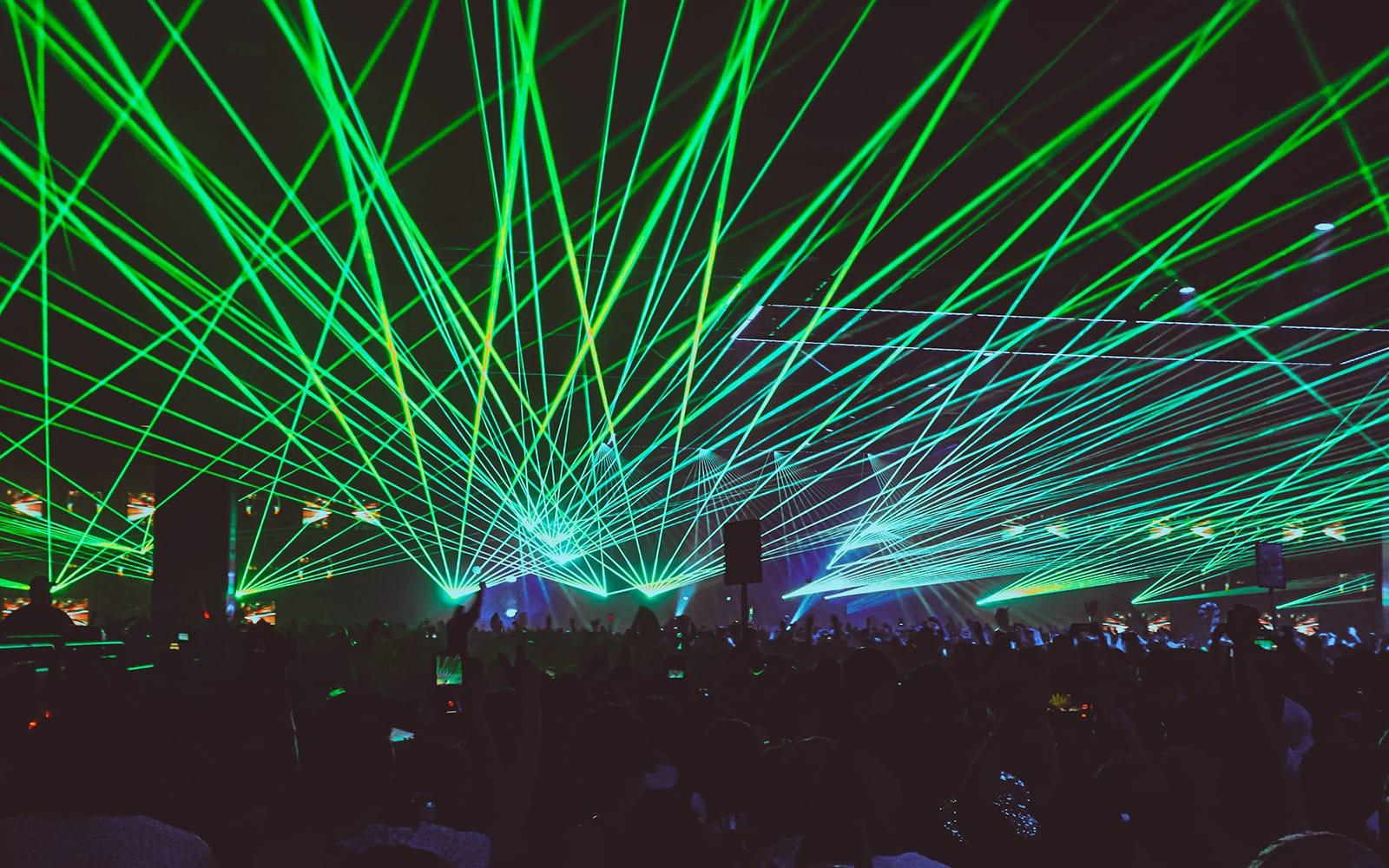A large crowd of people dancing at a rave as green lasers shoot overhead