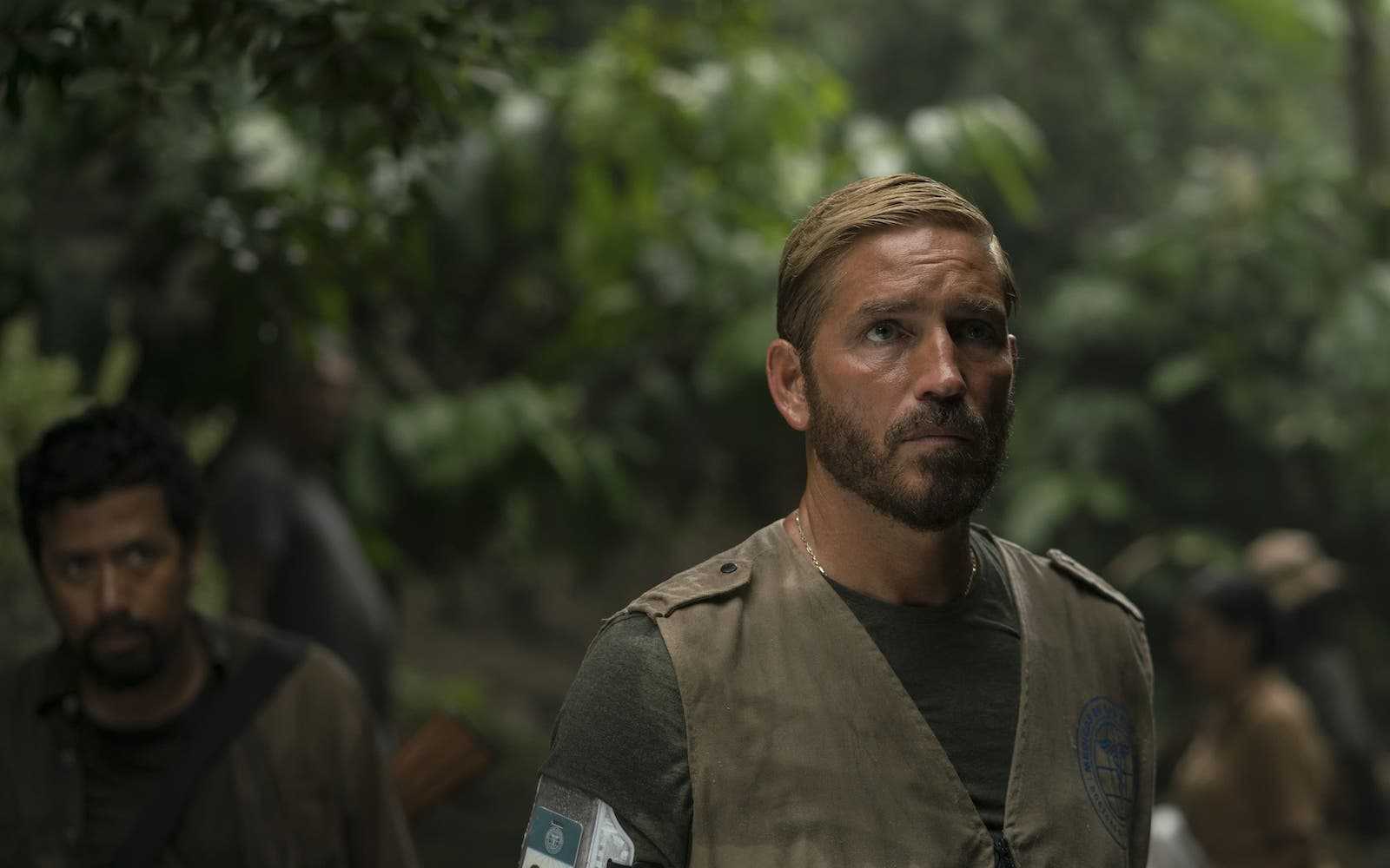 A man in camouflage stands in the jungle and stares off to the side with several people standing behind him