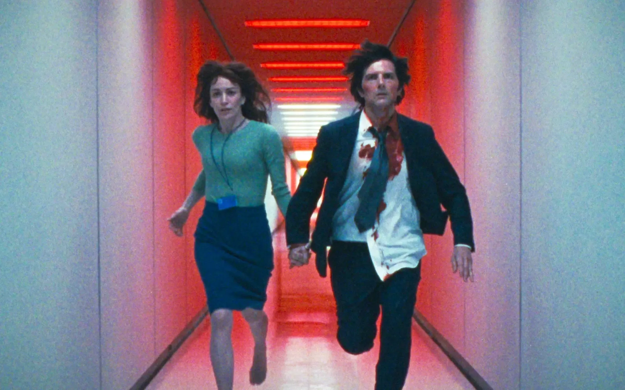 A man and woman run down a hallway while holding hands as red warning lights flash around them. They have worried looks on their faces and the man is covered in blood.