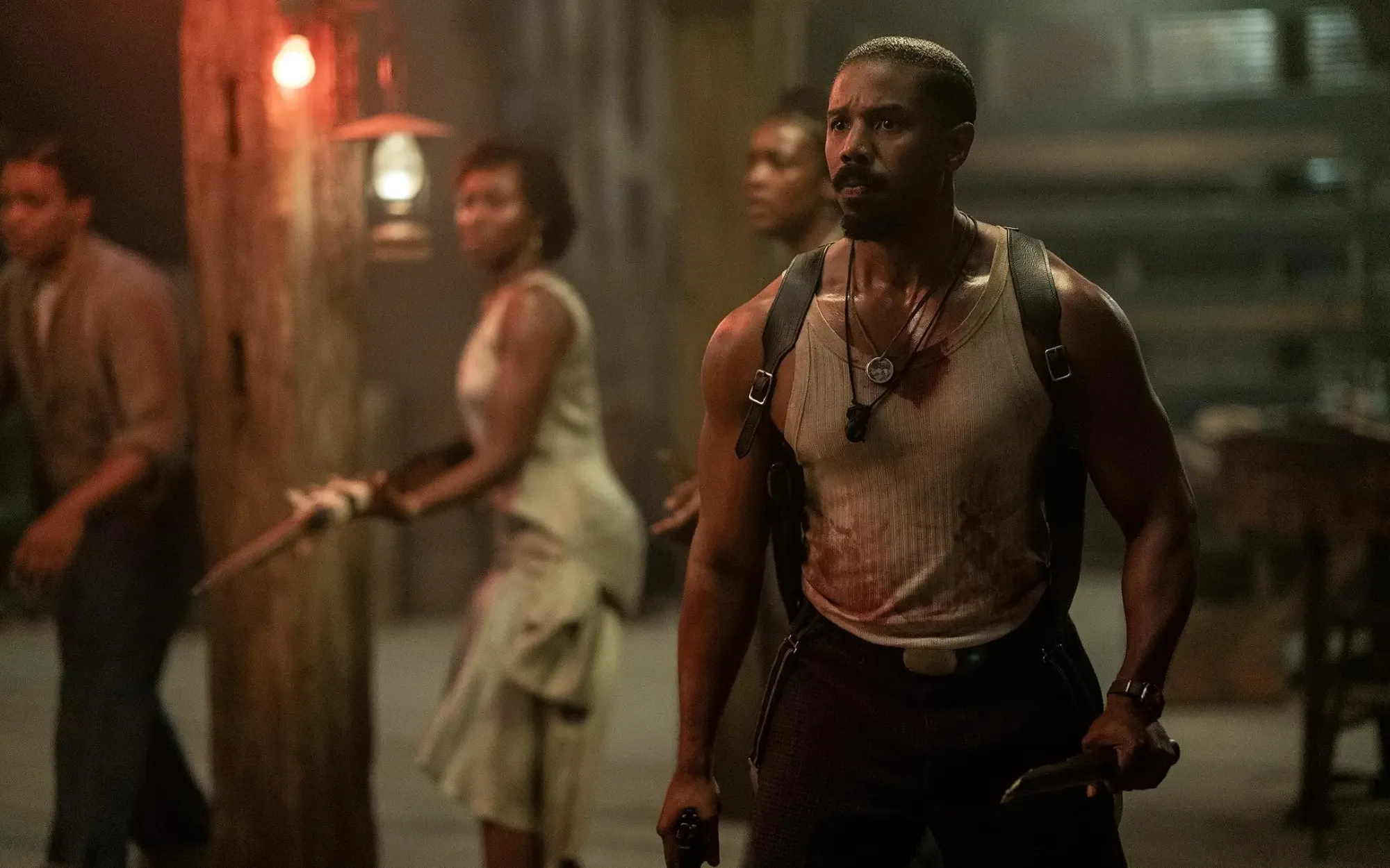 A Black man in a bloody shirt carrying a gun and knife in his hands stares at something with a concerned expression. Several people stand behind him, carrying weapons, too.